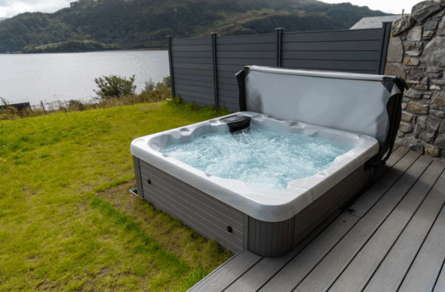A gray hot tub is sat on a gray decking, next to grass. In the background is a lake.