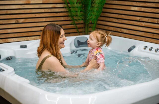 A mother and daughter sat in a white, hot tub. The hot tub is turned on. The background is made up of wood.