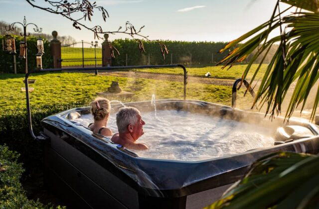 A couple are sat relaxing in a hot tub. In the background is a garden and a large garden hedge.
