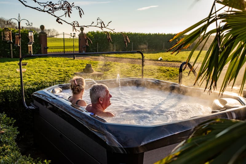 A couple are sat relaxing in a hot tub. In the background is a garden and a large garden hedge.